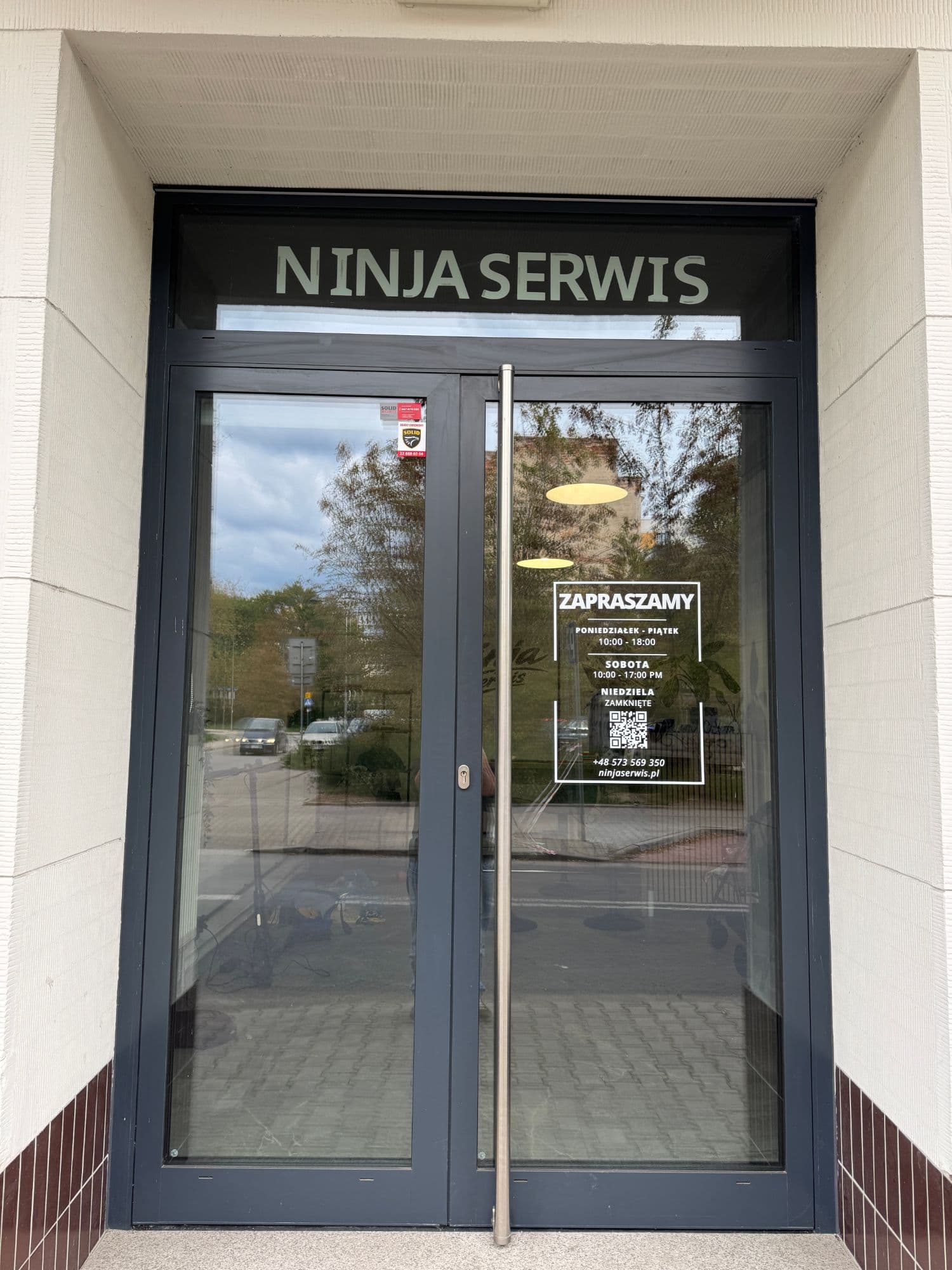 Wejście do serwisu Ninja Serwis przy ul. Folwarcznej 7/1A w Warszawie — przeszklone drzwi z szyldem i godzinami otwarcia