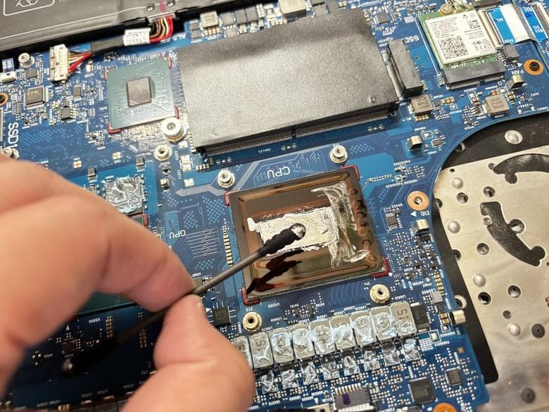 Nakładanie ciekłego metalu na odkryty rdzeń procesora w laptopie — płyta główna z widocznymi gniazdami pamięci i modułem Wi-Fi